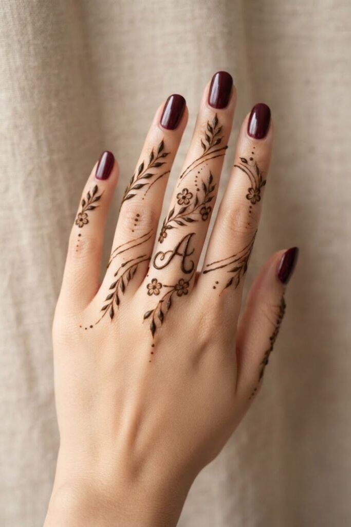 A letter finger mehndi design with delicate floral details and deep maroon nail polish